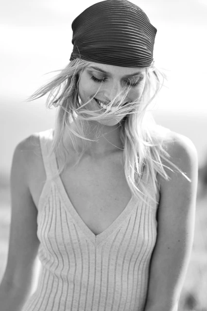 Black and white portrait of a young woman smiling in a sleeveless shirt and a headscarf with her blonde hair blowing in the wind.