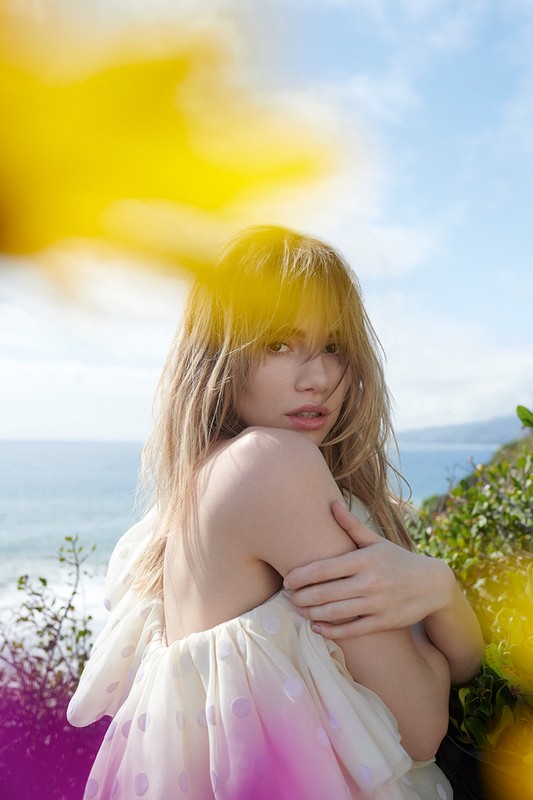 A young, fair-skinned woman with blonde hair poses outdoors against a blurred landscape with diffused yellow light in the foreground.