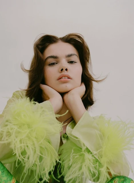 Portrait of a young woman with brown hair resting her chin on her hands, wearing a green blouse with feathered sleeves against a light background.