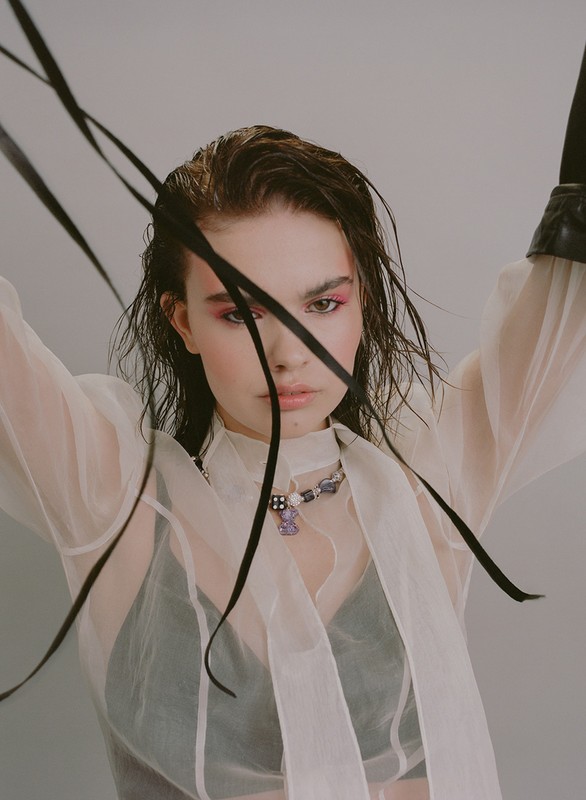 Close-up of a young woman wearing a sheer white blouse, a beaded necklace, and pink eyeshadow, with black ribbons framing her face.