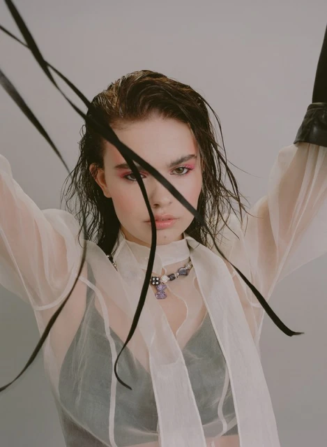 Close-up of a young woman wearing a sheer white blouse, a beaded necklace, and pink eyeshadow, with black ribbons framing her face.