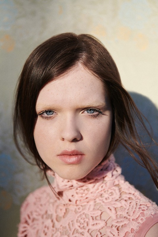 Portrait of a young woman with light skin and dark hair wearing a pink lace top against a textured yellow and blue wall.