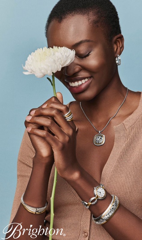 Black woman with short hair holding a white flower in front of her face, wearing jewellery and a brown ribbed top.