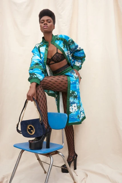 A woman in a tropical-themed outfit poses on a chair with a purse and platform heels against a neutral backdrop in a fashion setting.
