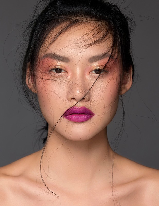 Close-up portrait of a young Asian woman with colorful makeup and loose hair, set against a dark grey background in a beauty shot.