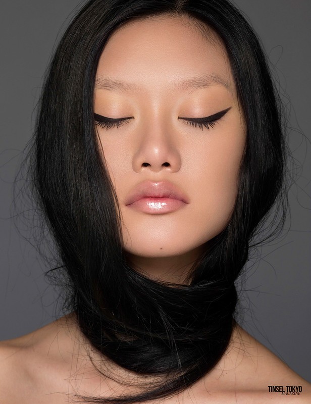 Close-up of an East Asian woman with long black hair draped around her neck, eyes closed, and glossy lips against a grey background.