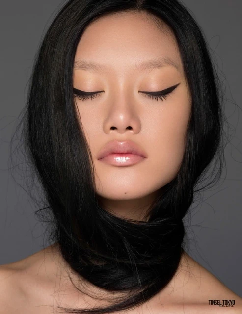 Close-up of an East Asian woman with long black hair draped around her neck, eyes closed, and glossy lips against a grey background.