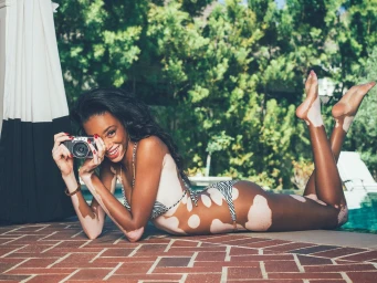 Model Winnie Harlow lying prone by a pool holding a camera, wearing a zebra print bikini with makeup by Simon Rihana.