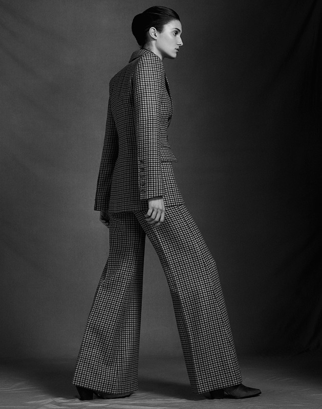 Monochrome portrait of a model standing in profile wearing a houndstooth blazer and flared trousers with hair styled in an updo.