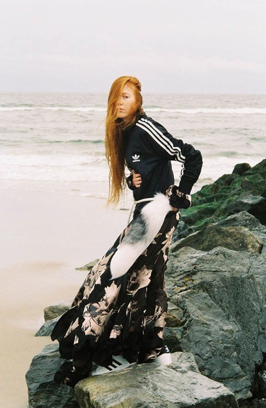 A person stands on rocks at a beach wearing a black Adidas jacket, and floral maxi skirt, facing camera. Cloudy sky and ocean backdrop.