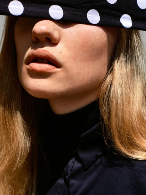 Close-up portrait of a blonde woman with a polka dot headscarf covering her eyes, wearing a black turtleneck shirt against a neutral background.
