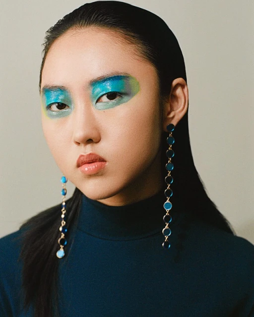 Close-up of Asian female model with vibrant blue and green eyeshadow, wearing a dark blue top and long blue dangling earrings.