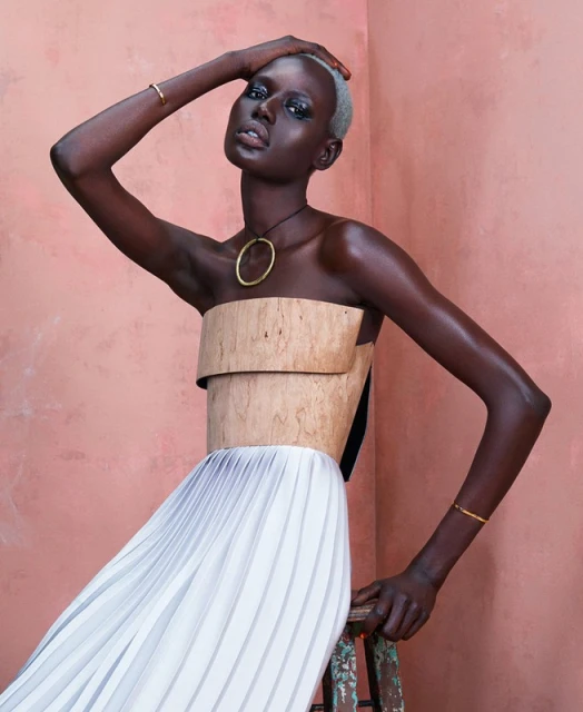 Fashion shot of a slender young woman with a shaved head wearing a two-piece top and pleated skirt in a photo studio against a coral backdrop.