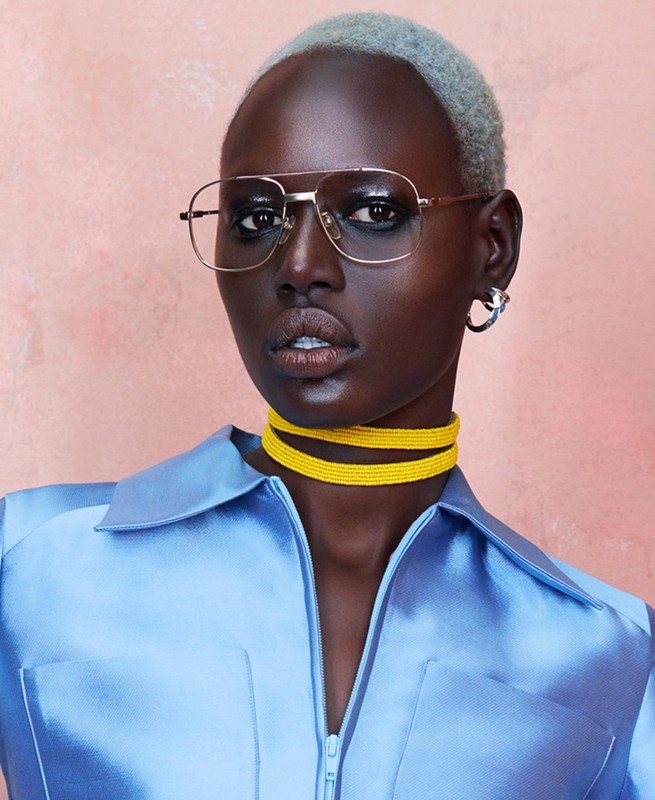 Close-up of a dark-skinned adult woman wearing glasses and a blue satin shirt against a mottled peach background in a studio setting.