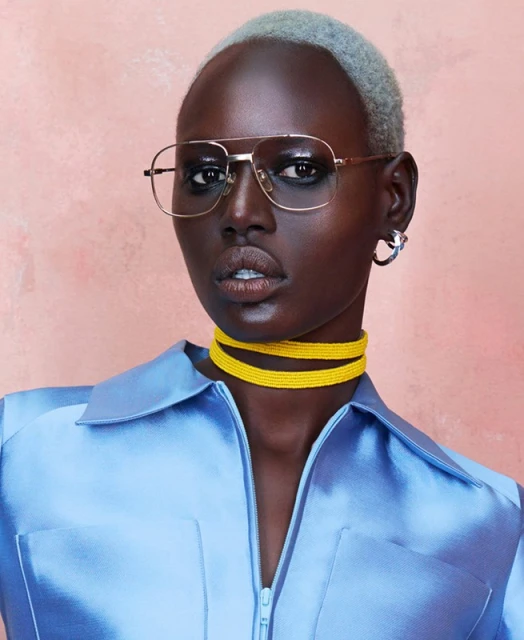 Close-up of a dark-skinned adult woman wearing glasses and a blue satin shirt against a mottled peach background in a studio setting.