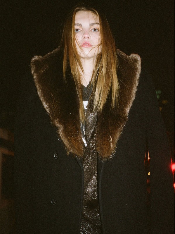 Woman in a dark coat with a large fur collar stands outdoors at night with soft lighting on her face and upper body in an urban environment.