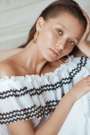 Close-up portrait of a woman with wet hair wearing a white off-the-shoulder dress with black zigzag trim and gold hoop earrings.