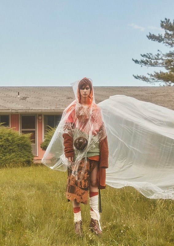 Adult fashion model poses outdoors in a clear plastic cape and layered clothing in front of a house and a large, translucent plastic sheet.
