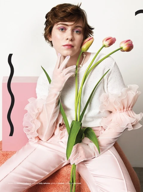 Portrait of a young woman with short brown hair, wearing a white sweater with sheer pink sleeves, and holding pink tulips.