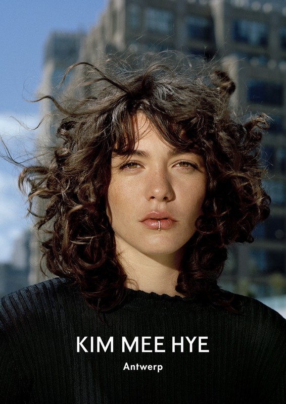 Close-up portrait of a young woman with curly brown hair and a lip ring, posing outdoors against a backdrop of tall buildings.