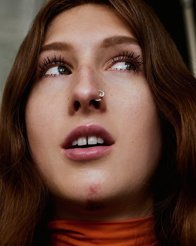 Low angle close-up of a young woman with auburn hair, fair skin, nose ring, wearing an orange top, looking away from the camera.