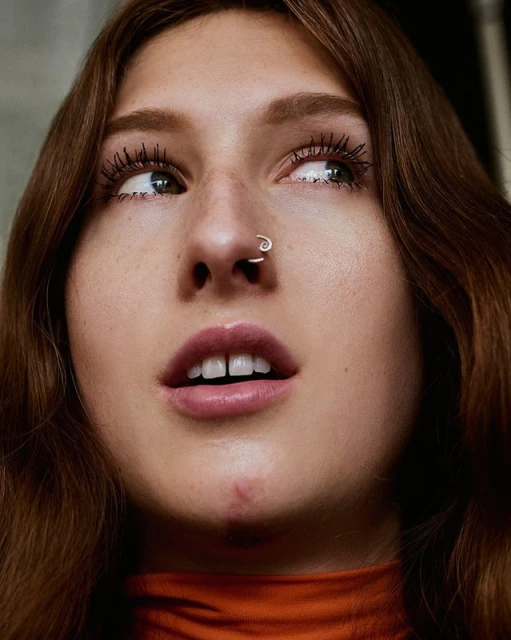 Low angle close-up of a young woman with auburn hair, fair skin, nose ring, wearing an orange top, looking away from the camera.