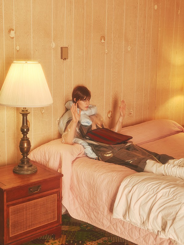 A woman in a transparent raincoat lies on a pink bed in a vintage-styled room with patterned wallpaper and a wooden bedside table.