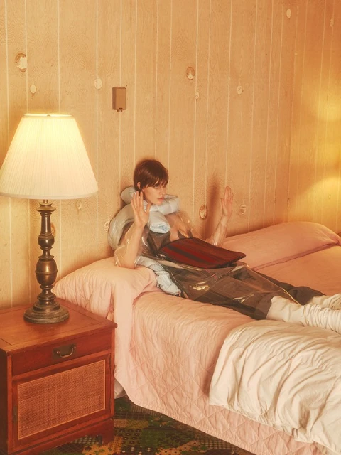 A woman in a transparent raincoat lies on a pink bed in a vintage-styled room with patterned wallpaper and a wooden bedside table.