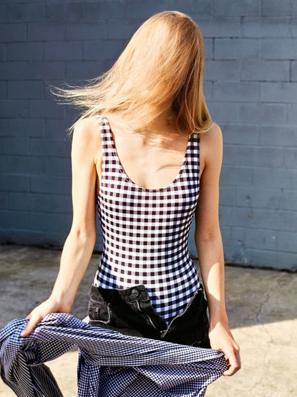 Blonde woman in a checkered swimsuit stands against a brick wall holding a denim shirt and open denim shorts in an outdoor setting.