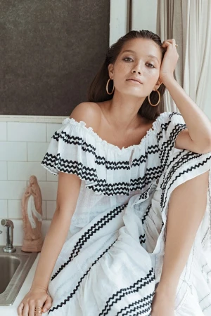 Woman with wet hair and hoop earrings wearing a white off-the-shoulder dress with black zigzag patterns