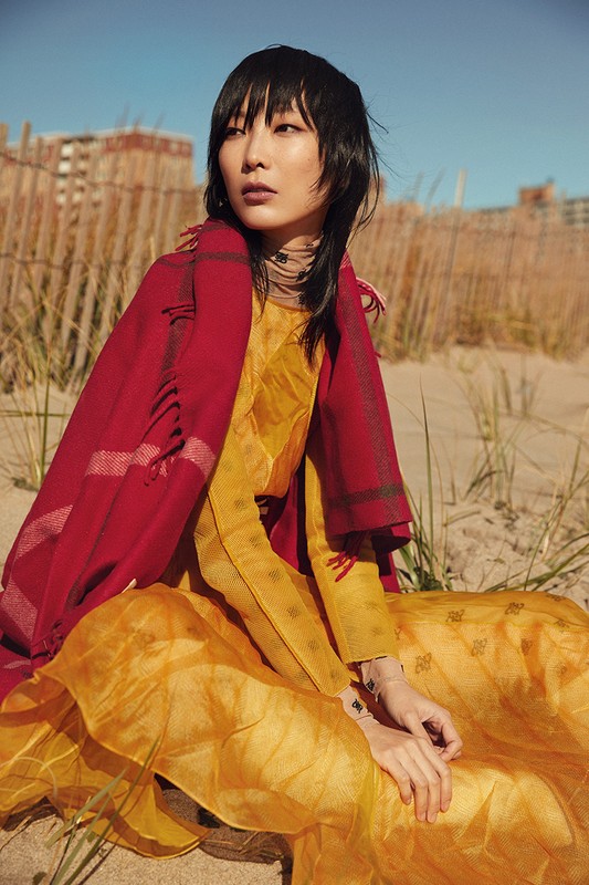 A young Asian woman in a yellow dress sits on a sandy beach, draped with a red shawl, gazing away with a serene expression.