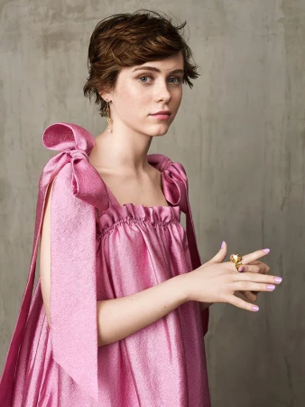 Portrait of a woman with short brown hair wearing a shiny pink dress with bow straps and a gold ring against a textured wall
