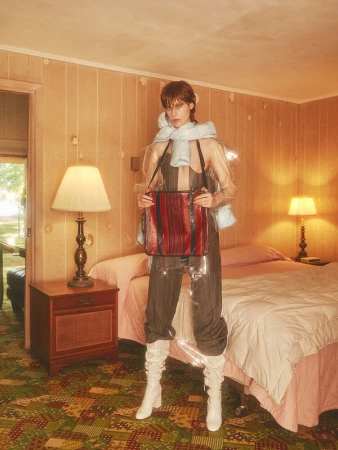 Fashionable woman poses in a vintage room wearing a transparent vinyl coat, gray striped pants, white boots, and holding a leather bag.