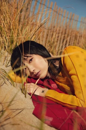 Woman lying on sand near tall grass wearing a yellow top and red blanket with hair styled by Sirsa