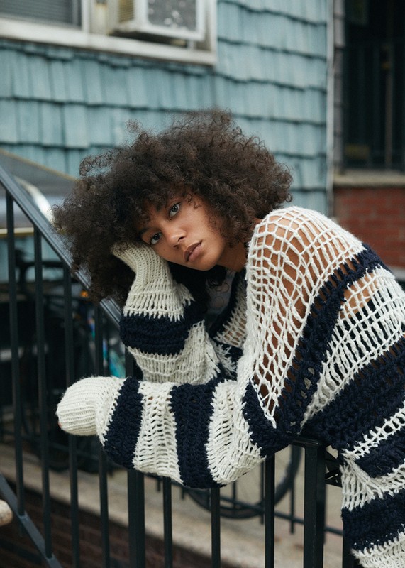 Young woman with afro hairstyle in a striped knit sweater leans on an iron railing with a blue-green paneled exterior wall in the background.