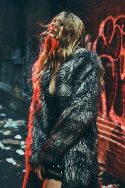 Blonde woman in a fur coat stands against a brick wall with graffiti. She is illuminated by dramatic red and orange lighting.