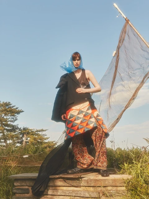 A woman stands on stone steps, wearing a blazer, print pants, and a blue headscarf, with a large net structure behind her outdoors.