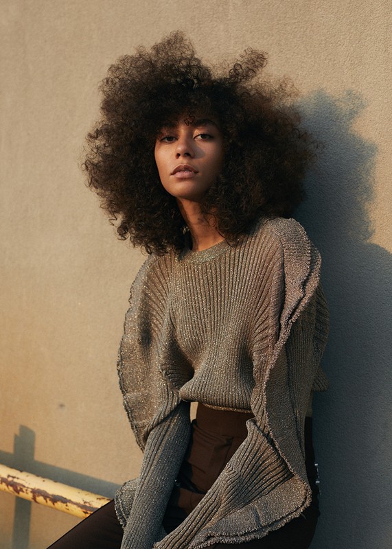Portrait of a woman with natural afro hair wearing a metallic, ruffled sweater in front of a textured concrete wall.
