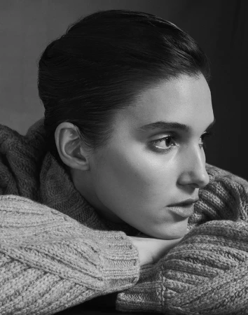 Close-up profile portrait of a woman with braided hair wearing a chunky knit sweater resting her chin on her crossed arms.