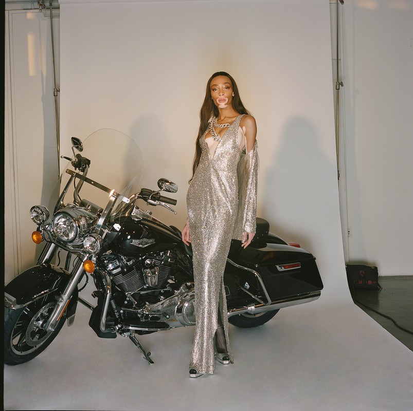 Winnie Harlow poses next to a black Harley Davidson motorcycle in a studio setting, wearing a long, silver, sparkling gown.