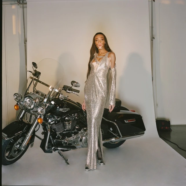 Winnie Harlow poses next to a black Harley Davidson motorcycle in a studio setting, wearing a long, silver, sparkling gown.