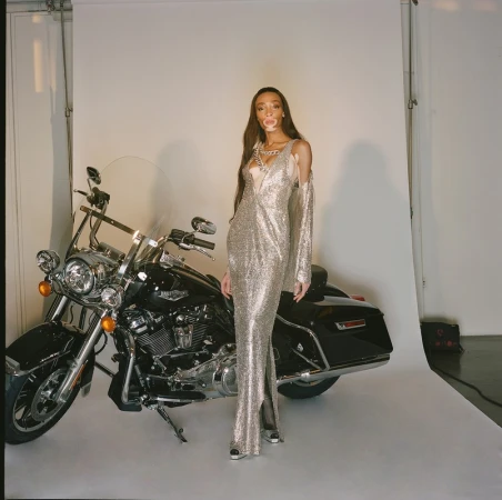Winnie Harlow poses next to a black Harley Davidson motorcycle in a studio setting, wearing a long, silver, sparkling gown.