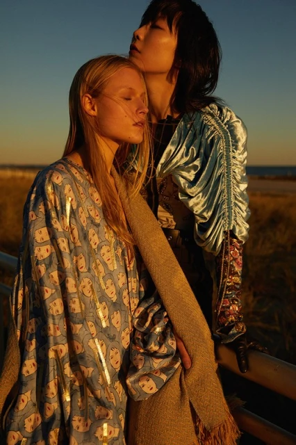 Two women pose outdoors at golden hour. One woman with blonde hair wears a face-print dress; the other has black hair and a blue top.