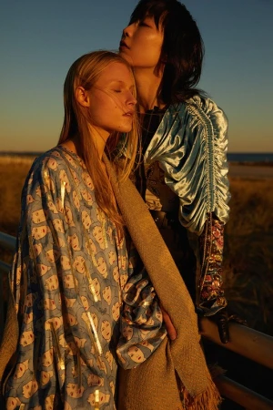 Two women pose outdoors at golden hour. One woman with blonde hair wears a face-print dress; the other has black hair and a blue top.