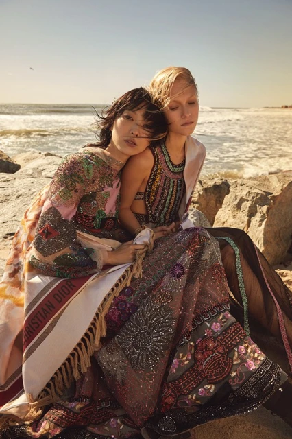 Two fashion models in embellished gowns sit on a rocky beach with ocean waves in the background. Includes a Christian Dior blanket.