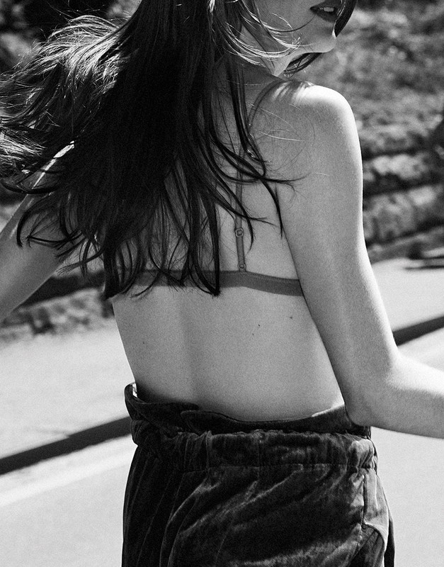 Monochrome close-up of a woman in motion with long dark hair blowing, wearing a thin-strap top and dark trousers outdoors.