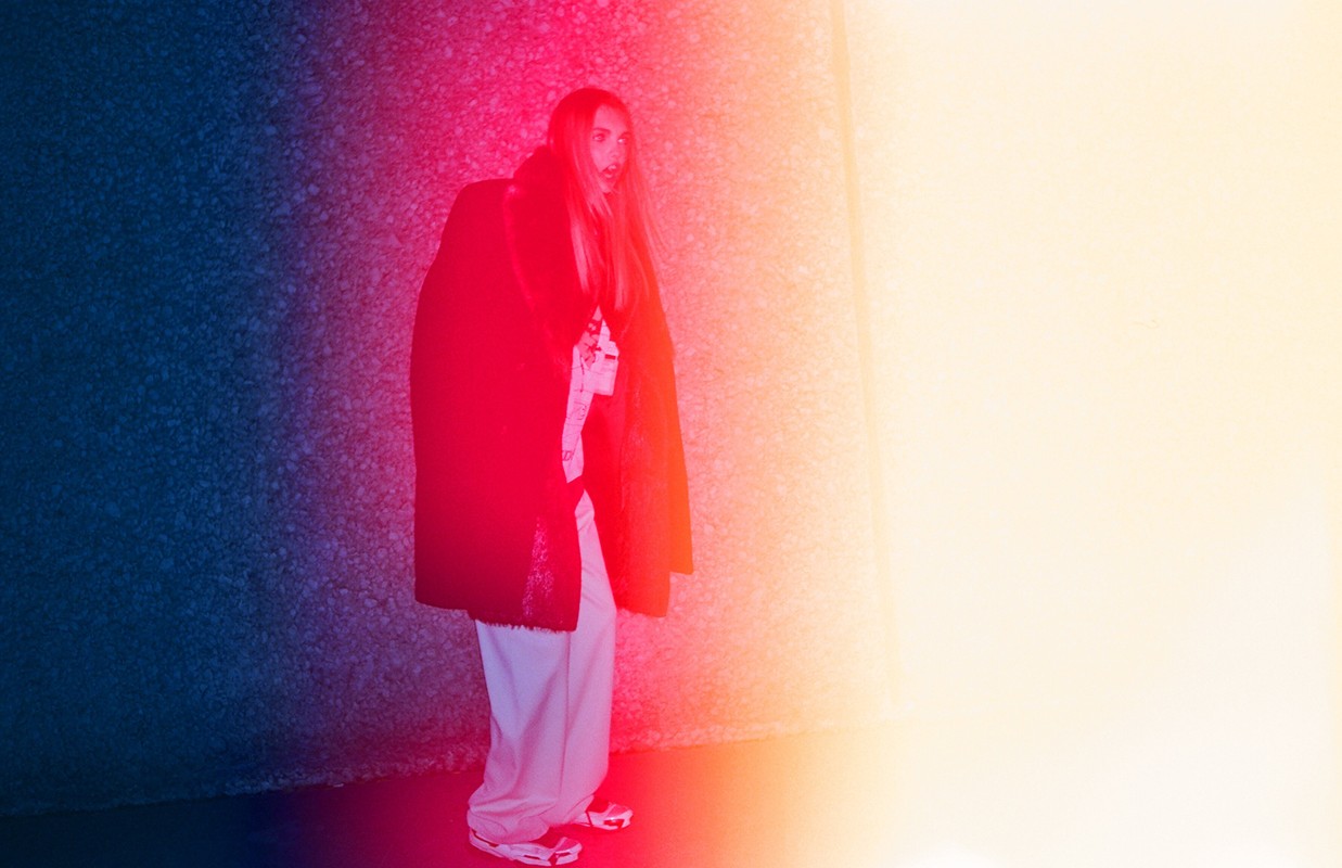 Model with long blonde hair wearing a dark coat and light trousers bathed in red, orange, and blue lighting in Los Angeles.