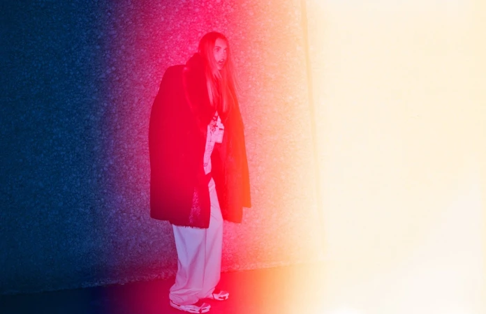 Model with long blonde hair wearing a dark coat and light trousers bathed in red, orange, and blue lighting in Los Angeles.