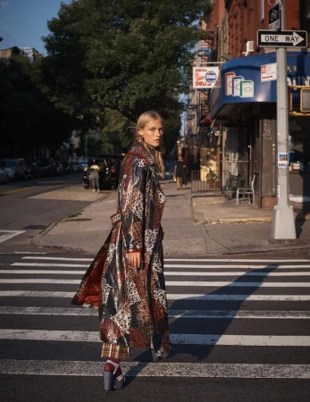 A fair-skinned fashion model in a patterned coat walks across a crosswalk in an urban street setting during daylight hours.