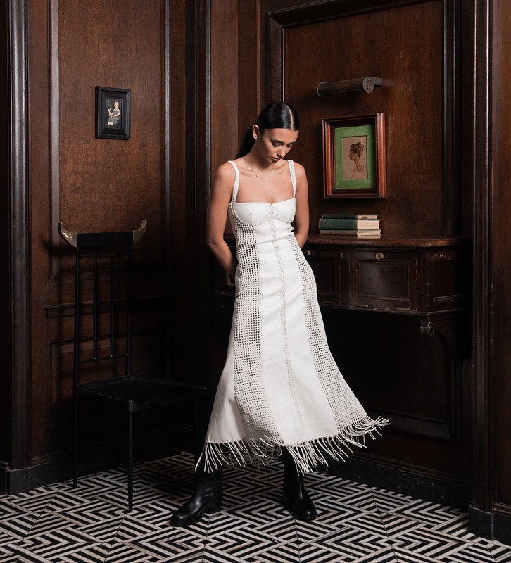 Adult female in a white dress poses indoors against dark wood paneling with a geometric tile floor and ornate chair and artwork.